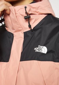 Veste rose clair et noire avec une fermeture éclair à l'avant. Dispose d'un col montant, d'un cordon de serrage ajustable et d'un logo sur la poitrine. Tissu texturé.