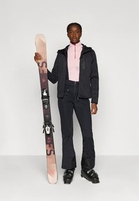 Czarna kurtka narciarska z różowym topem na zamek, czarne spodnie narciarskie, trzyma jasnokolorowe narty z czarnymi wiązaniami i nosi czarne buty narciarskie.