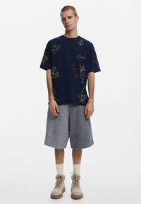 Marineblaues T-Shirt mit bunten gestickten Mustern, kombiniert mit grauen gestreiften Shorts. Tanfarbene Stiefel und weiße Socken runden den Look ab.