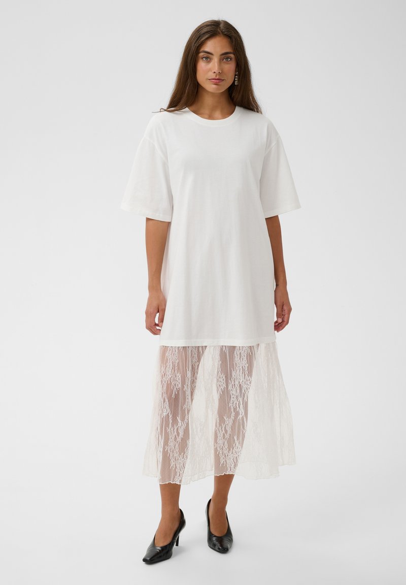 Femme aux longs cheveux bruns portant une robe blanche oversize avec des manches courtes et une partie inférieure en dentelle transparente, associée à des talons noirs.