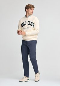 Felpa crema con la scritta "POLO CLUB" in testo verde scuro, abbinata a pantaloni blu navy e scarpe da ginnastica beige con accenti verdi.