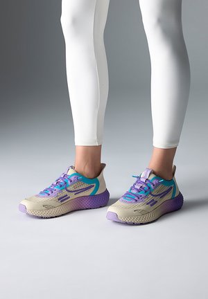 Füße, die beige, lila und türkisfarbene Laufschuhe mit strukturierten Sohlen tragen, kombiniert mit weißen, enganliegenden Leggings vor einem grauen Hintergrund.