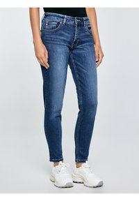 Nő középderékig érő kék skinny farmerben és fehér masszív tornacipőben, világos, egyszerű háttér előtt állva.