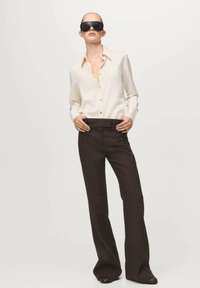 Donna con occhiali da sole neri oversize, camicetta color crema con bottoni e pantaloni a gamba larga a vita alta di colore marrone scuro, davanti a uno sfondo semplice.