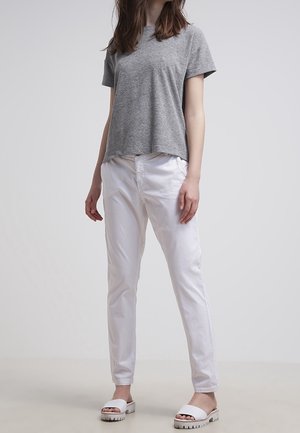 Osoba ubrana w szarą koszulkę z krótkim rękawem, białe spodnie i białe klapki, stojąca na jednolitym jasnym tle.