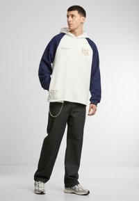 Sudadera con capucha de mangas raglán azul marino y cuerpo blanco, con acentos bordados en dorado. Combinada con jeans oscuros y zapatillas.