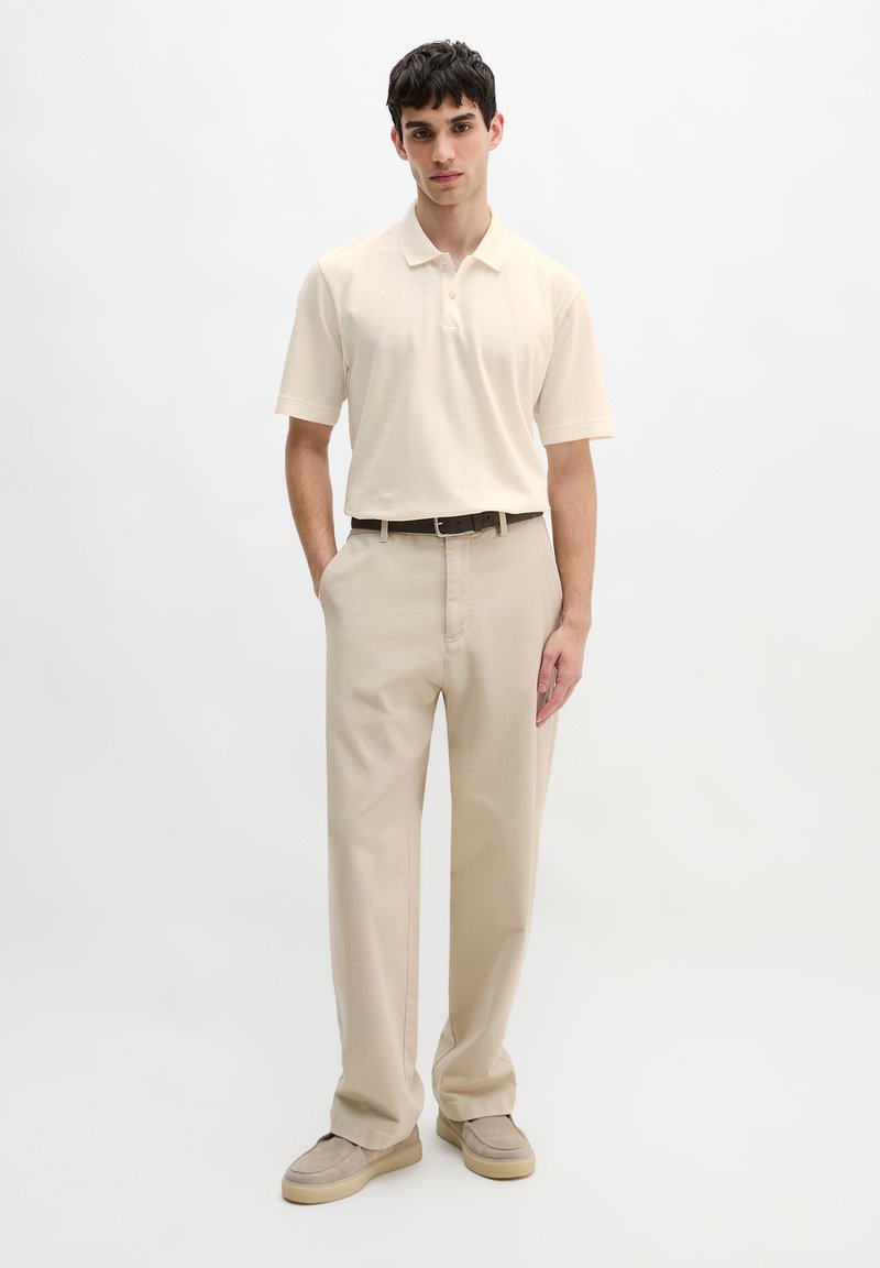 Junger Mann steht mit den Händen in den Taschen, trägt ein cremefarbenes kurzärmeliges Polo, beige Hosen, einen braunen Gürtel und beige Schuhe vor weißem Hintergrund.
