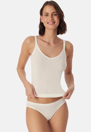 Donna che sorride indossando una canottiera di cotone bianco e biancheria intima abbinata, mentre sistema l'orlo della canottiera su uno sfondo neutro.