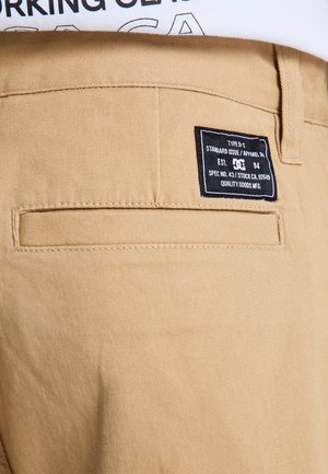 Beżowe bawełniane spodnie o fakturowanym wykończeniu, z czarną metką na tylnej kieszeni, zawierającą informacje o pielęgnacji i marce.
