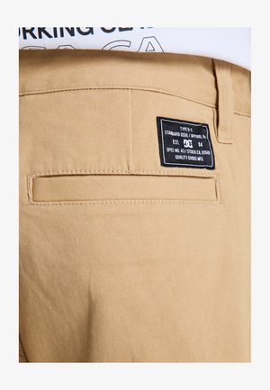 Beżowe bawełniane spodnie o fakturowanym wykończeniu, z czarną metką na tylnej kieszeni, zawierającą informacje o pielęgnacji i marce.