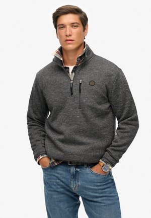 Sudadera de forro polar gris con cuello con cremallera, que cuenta con un bolsillo en el pecho y un pequeño logo. Lleva puesta sobre una camisa de cuadros y combinada con jeans azules.