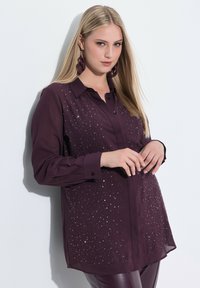 Tumman violetin sävyinen kauluspaidan, jossa pitkähihaisuus ja etuosassa hajallaan kevyesti heijastavia koristeita.