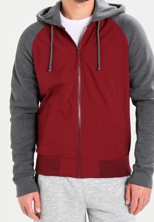 Mann trägt eine rot-graue Kapuzenjacke mit Reißverschluss und Kordeln, kombiniert mit hellgrauen Jogginghosen.