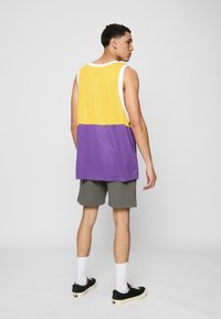 Flerfarget tanktopp med et gult og lilla design, laget av mesh-stoff. Kombinert med grå shorts og svarte joggesko. Hvite sokker synlige.