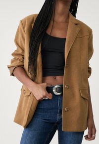 Blazer marron à coupe ajustée, tissu texturé, deux poches avant, et boutons contrastants, porté avec un haut noir et un jean bleu.