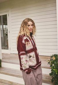 Gestrickter burgunderroter Pullover mit Blumenmustern, Reißverschluss vorne und strukturierter Oberfläche, kombiniert mit karierten Hosen, vor einem weißen Holzpaneel.
