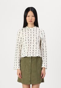 Valge kudumista blouse musta geomeetrilise mustriga, pika varrukaga ja lainedega alläärega, koos rohelise nahast nööpidega seelikuga.