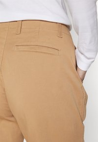 Vue arrière d'une personne portant un pantalon beige et une chemise blanche à manches longues, avec la main droite placée dans la poche latérale du pantalon.