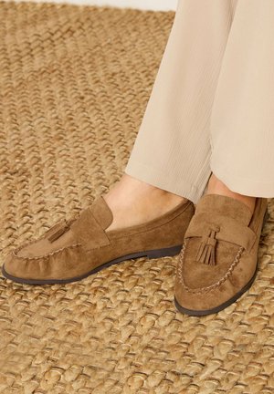 Bruine suède loafers met tassels gedragen met lichtbeige textuurbroek op een geweven tapijt van natuurlijke vezels.