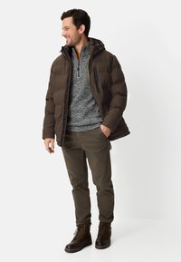 Mørkebrun dunjakke med hætte, grå tekstureret sweater, olivengrønne bukser og brune snørestøvler, stående mod en hvid baggrund.