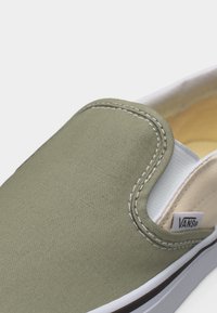 Slip-on παπούτσι Vans σε ελιά πράσινο καμβά με λευκό ελαστικό πλευρικό πάνελ, λευκή ραφή και λαστιχένια σόλα με ετικέτα λογότυπου Vans.