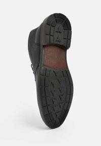 Calzado negro con suela de goma que presenta un patrón texturizado, forma redondeada y un acento marrón en el talón.