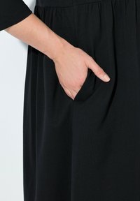 Černé šaty vyrobené z měkké látky, s nařaseným pasem a boční kapsou, hladkým povrchem a rukávy do poloviny délky.