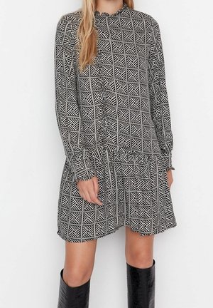 Kobieta w czarno-białej sukience z długim rękawem o geometrycznym wzorze, w połączeniu z czarnymi, teksturowanymi kozakami sięgającymi do kolan.