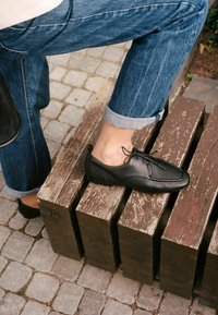 Personne portant un jean bleu retroussé et des chaussures à lacets en cuir noir, reposant un pied sur un banc en bois usé sur une surface pavée.