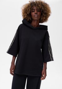 Czarna, oversize bluza z kapturem, z krótkimi rękawami i zamkami błyskawicznymi na rękawach. Wykonana z miękkiego, fakturowanego materiału.