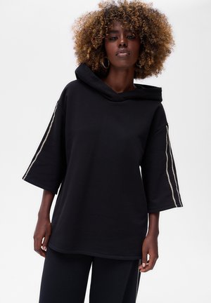 Haut à capuche noir oversize avec un col montant, des manches courtes et des accents zippés le long des manches. Fabriqué en tissu doux et texturé.