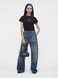 Γυναίκα που φοράει μαύρο t-shirt Margiela, μπλε τζιν με φαρδιά πόδια, μαύρα παπούτσια και κρατά μια μικρή μαύρη τσάντα με μακριές λαβές.