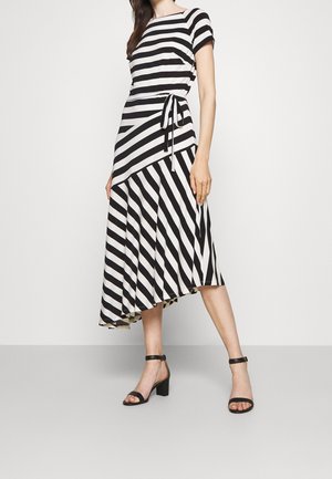 Žena v černobílých pruhovaných krátkých šatech s asymetrickým lemem a černých sandálech na podpatku s kotníkovým páskem stojí na šedé podlaze.