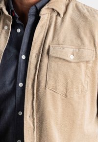 Beige corduroy overhemdjack met een licht gestructureerd oppervlak, voorzien van een borstzak en witte knoopsluitingen, gecombineerd met een donkerblauw shirt eronder.
