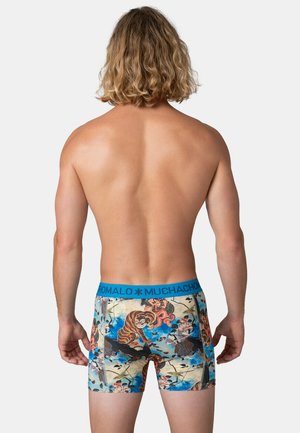 Man met schouderlang blond haar staat met de rug naar de kijker toe en draagt kleurrijke boxershorts met tijger- en vogelmotieven en blauwe tailleband met tekst.