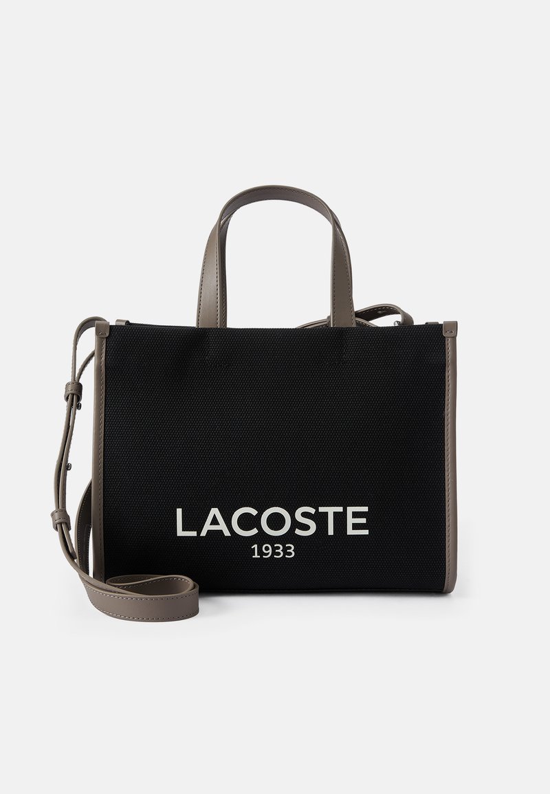 Bolsa de tela negra con detalles en cuero sintético gris, que presenta el logo "LACOSTE 1933" en blanco, asas dobles y correa de hombro desmontable.