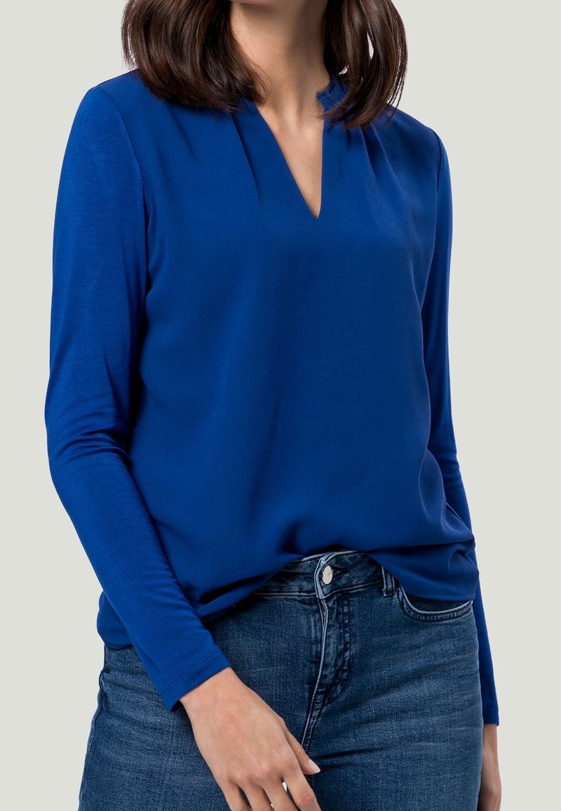 Donna che indossa una blouse a maniche lunghe di un blu reale con scollatura a V infilata in jeans blu scuro, su uno sfondo chiaro e semplice.