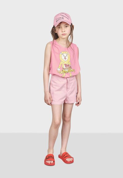 Camisa sin mangas rosa con un gráfico de león, pantalones cortos rosas y sandalias rojas con texto negro. Lleva una gorra rosa con texto estilizado.