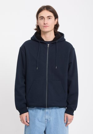 Jeune homme portant un sweat à capuche zippé bleu marine avec cordons et un jean bleu clair, debout devant un fond blanc uni.