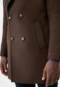 Cappotto in lana marrone con superficie strutturata, design doppiopetto, quattro bottoni in metallo e taschino laterale a sinistra.