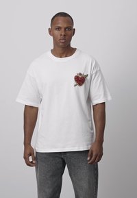 Weißes Baumwoll-T-Shirt mit kurzen Ärmeln, versehen mit einem roten, mit Schmucksteinen besetzten Herz- und Schwert-Design auf der linken Brustseite. Lockerer Schnitt, Rundhalsausschnitt.