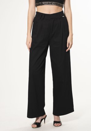 Zwarte wijdere broeken met plooien, hoge taille en een gladde textuur. Gecombineerd met zwarte open-toe hakken en een kort zwart topje met versieringen.