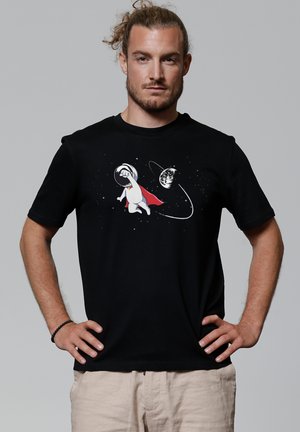 Zwarte katoenen t-shirt met een afbeelding van een astronautenhond met een rode cape, omringd door sterren en een planeet, op een effen achtergrond.