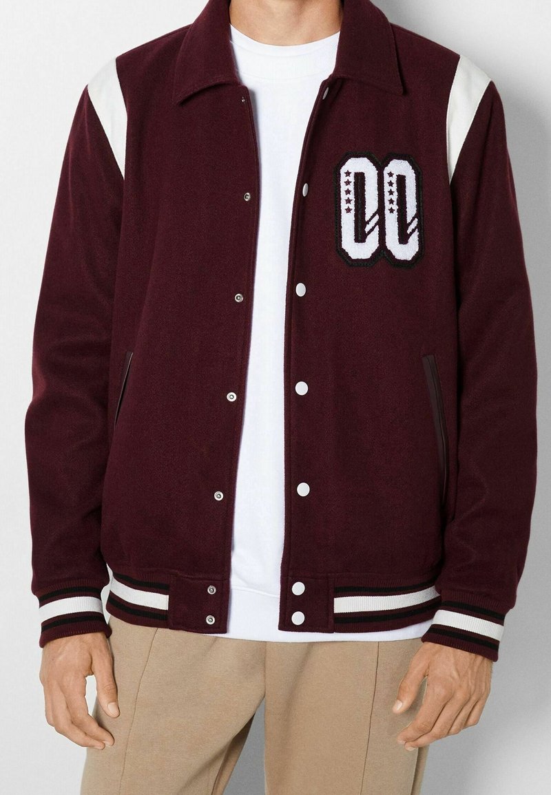 Homme portant une veste universitaire bordeaux avec des rayures blanches aux épaules, un grand patch "00" sur la poitrine, des boutons blancs et des poignets rayés.