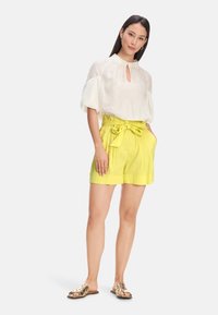 Weiße, locker sitzende Bluse mit Schlüsselaugen-Ausschnitt. Helle gelbe, hoch taillierte Shorts mit Schleifenbändchen und Taschen. Flache Sandalen mit goldfarbenen Akzenten.