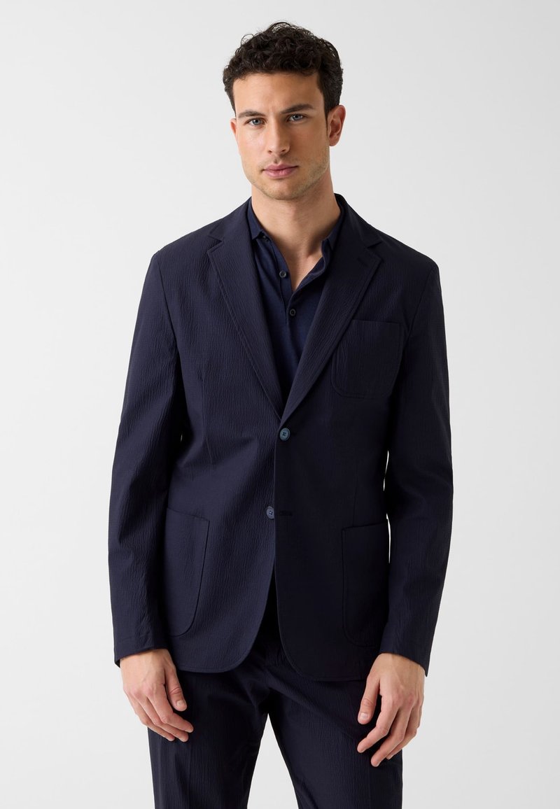 Homme aux cheveux courts et bouclés portant un blazer bleu marine foncé et un pantalon assorti, debout devant un fond clair uni.