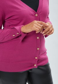 Cardigan fuchsia avec décolleté en V, poignets côtelés et boutons à carreaux dorés. Texture douce avec une coupe ajustée, associé à un pantalon noir.