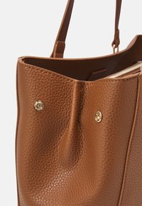 Hnědá strukturovaná kožená taška tote s ozdobami ze zlatého kovu a měkkým řaseným designem. Otevřený vršek s krátkými rukojetmi pro nošení.