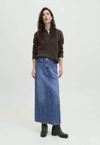 Suéter castanho de malha com gola, combinado com uma saia de ganga azul longa, com costuras verticais e bainha desfiada, complementado por botas pretas.