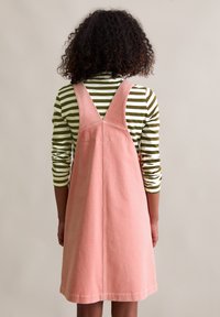 Ein Kind mit lockigem Haar, das ein rosa Cordkleid über einem grün-weiß gestreiften Langarmshirt trägt, sieht von der Kamera weg.